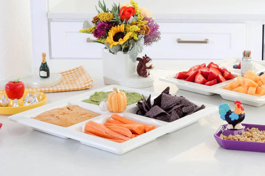 Nora Fleming sectional server with four compartments holding carrot sticks, chips, dip, and spread on a kitchen counter