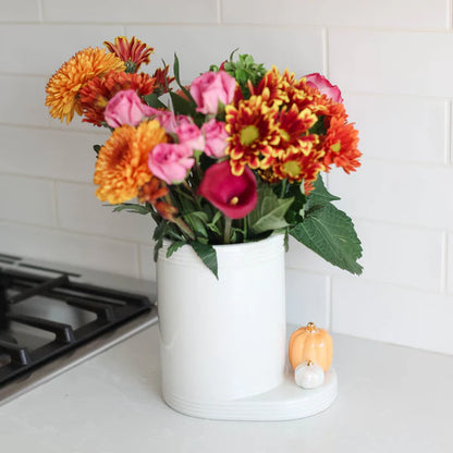 Nora Fleming utensil holder in white used as a vase with colorful flowers on kitchen counter